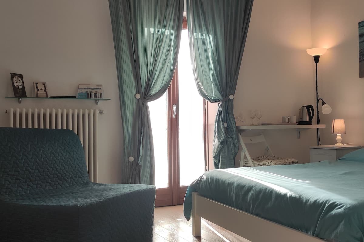 A bedroom with a bed, a chair, a radiator, a shelf with items, and a painting on the wall.