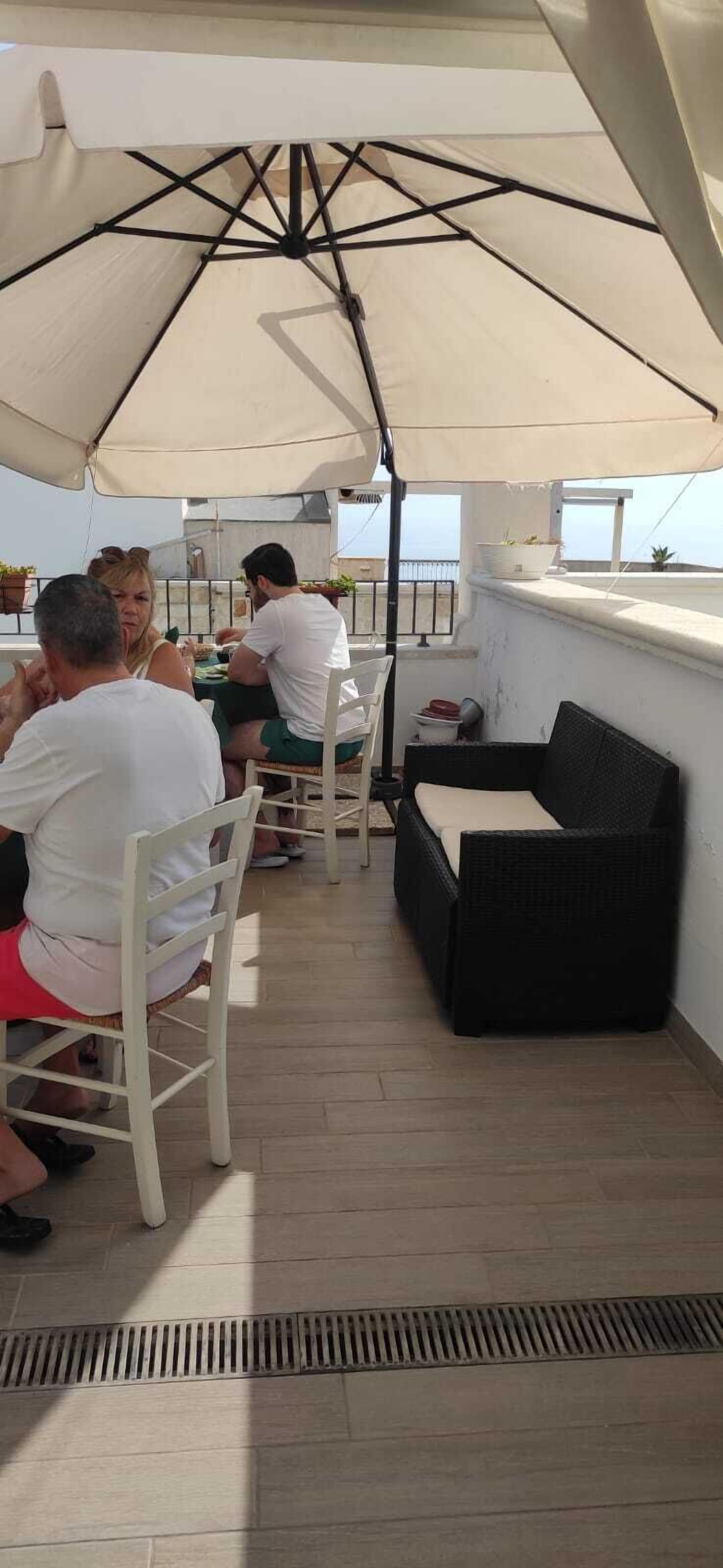 Ein überdachter Außenbereich mit Tischen und Stühlen, einem schwarzen Rattansofa und einem Blumentopf auf einem Balkon.