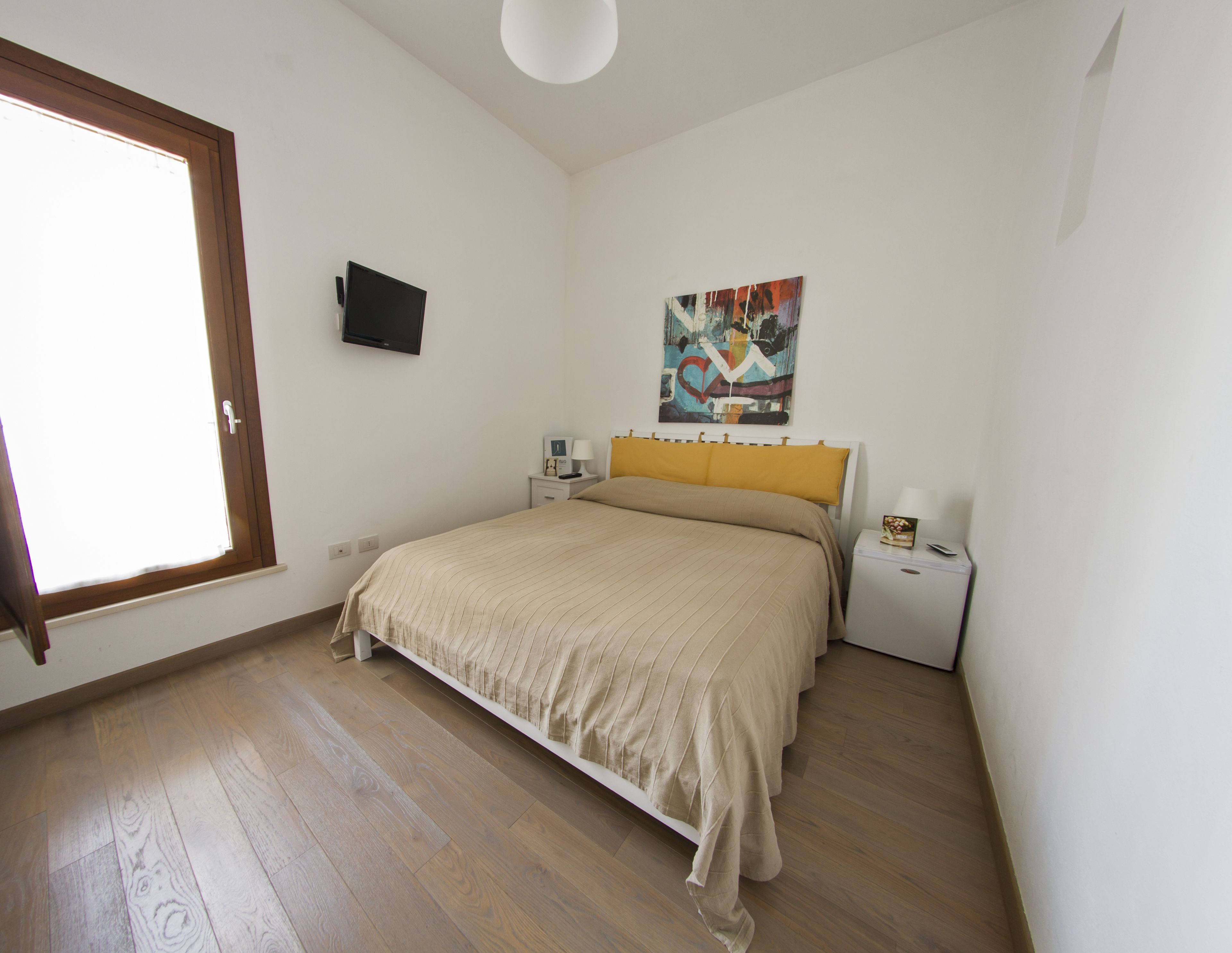 Un dormitorio con cama, mesitas de noche, televisión, un cuadro y ventana.