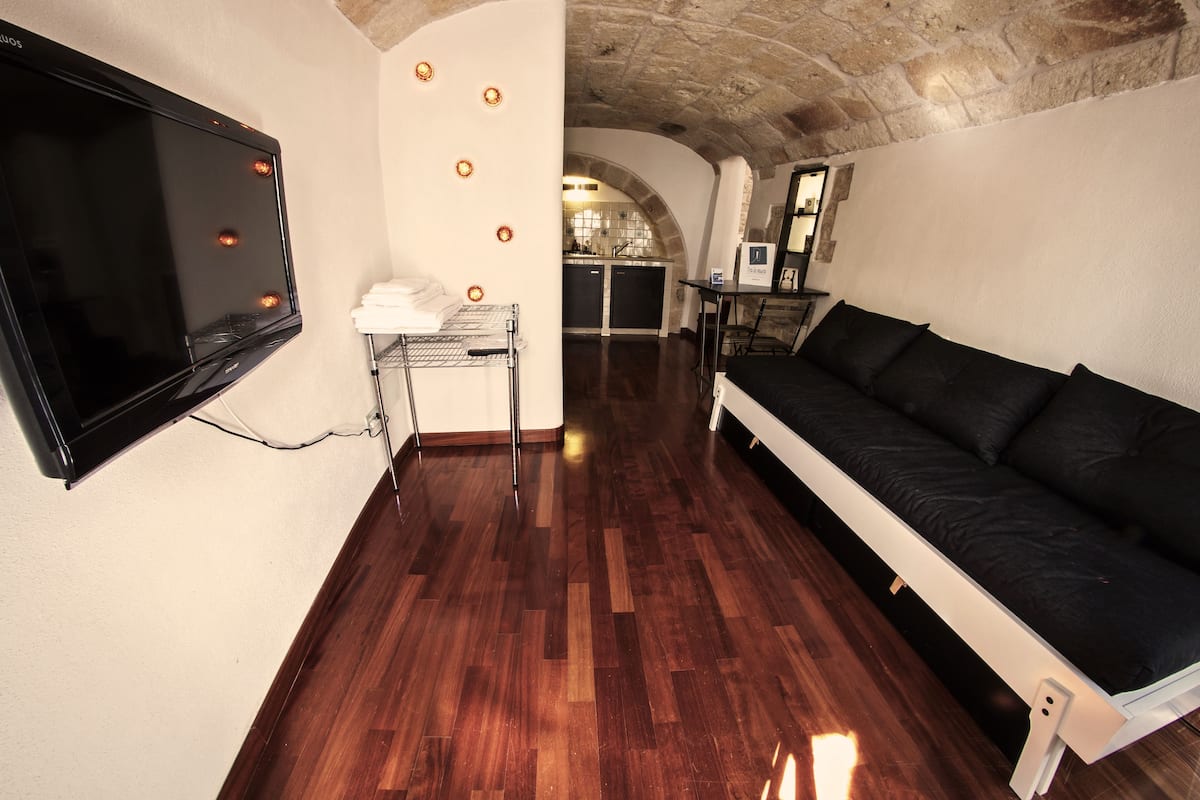 A room with a wooden floor, a black sofa, a flat-screen TV mounted on the wall, and a kitchen area in the background.