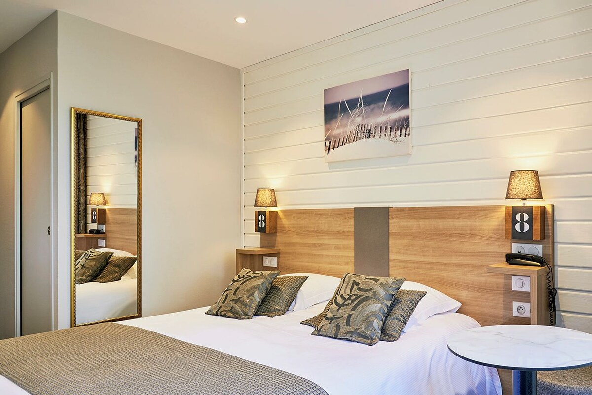 A hotel room with a bed, bedside lamps, a round table, a mirror, and a wall-mounted artwork.
