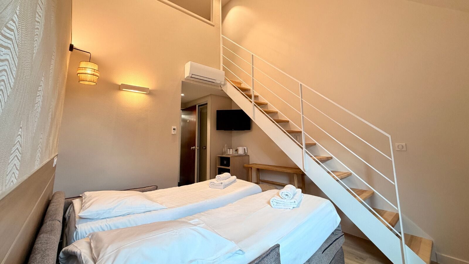 Habitación de hotel con dos camas, una escalera, un equipo de aire acondicionado y un televisor de pantalla plana montado en la pared.