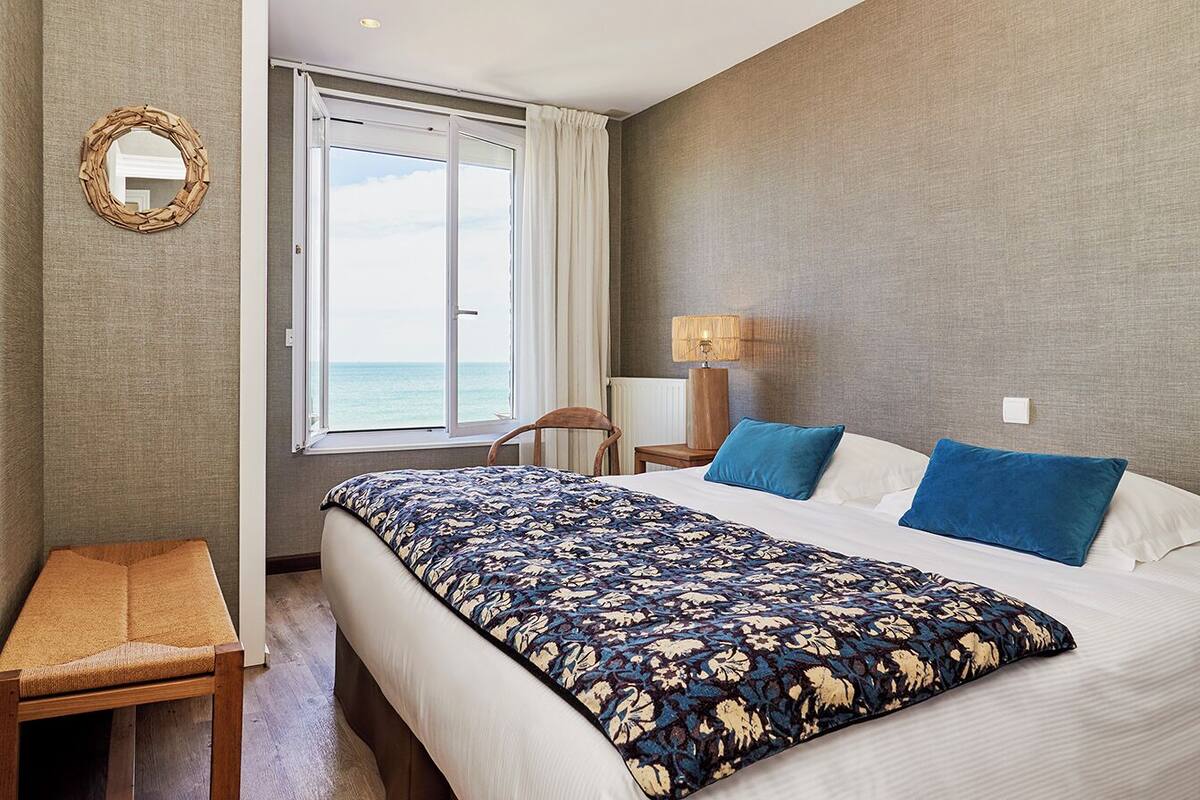 A hotel room with a bed, a bedside table, a chair, a lamp, and a view of the sea through a window.