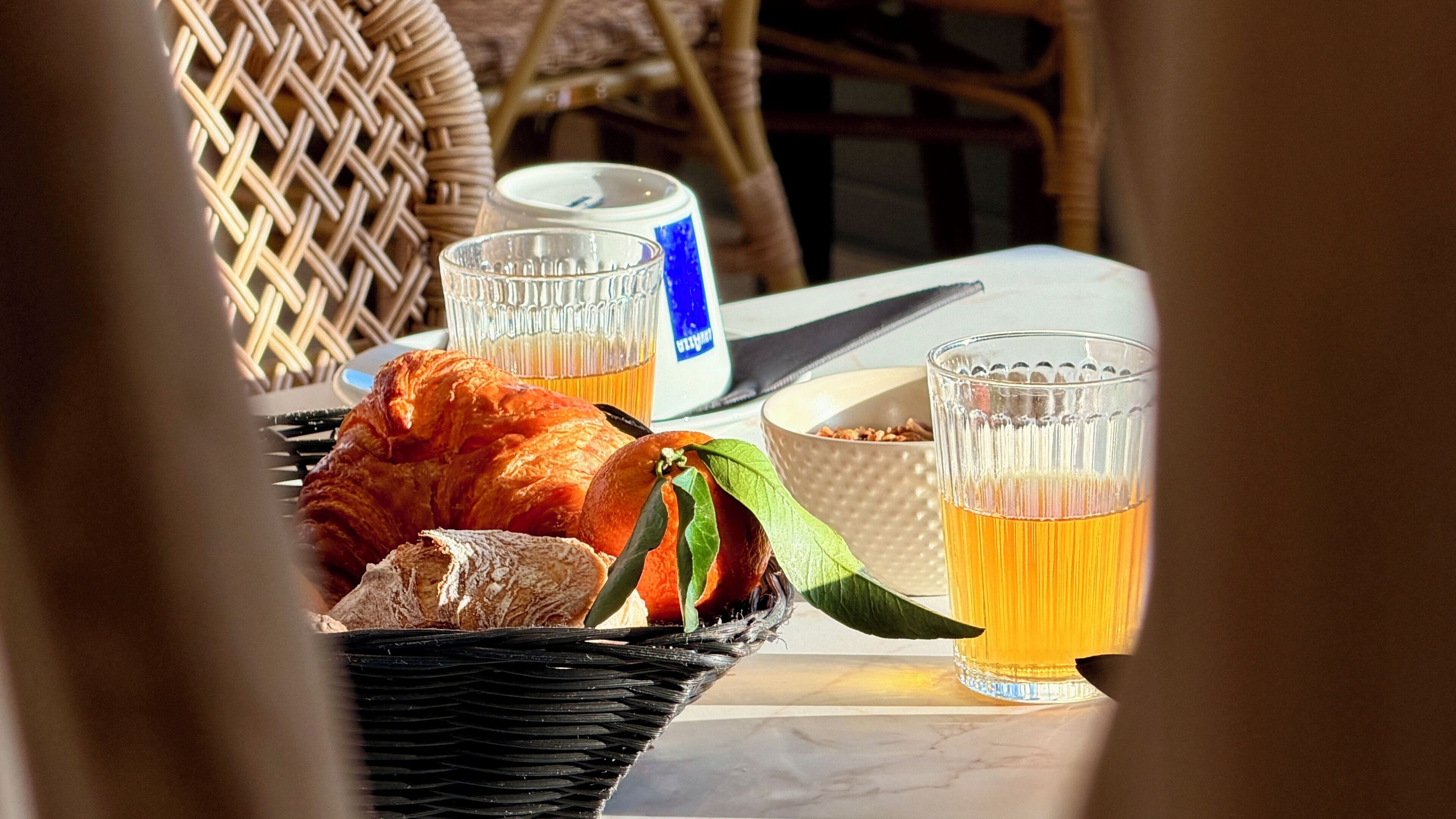 Una mesa con una cesta que contiene pan, un croissant y una fruta con hojas. Hay dos vasos de líquido ámbar, probablemente cerveza, y una botella con etiqueta azul.
