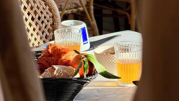 Una mesa con una cesta que contiene pan, un croissant y una fruta con hojas. Hay dos vasos de líquido ámbar, probablemente cerveza, y una botella con etiqueta azul.