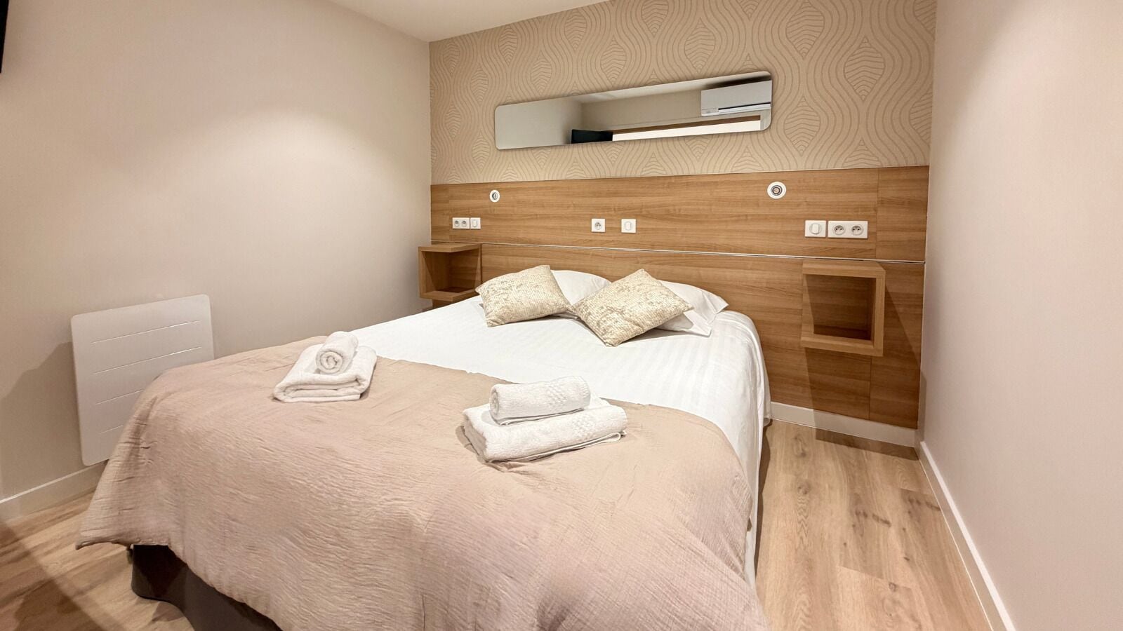 A neatly made bed with a beige blanket, two pillows, and rolled towels.