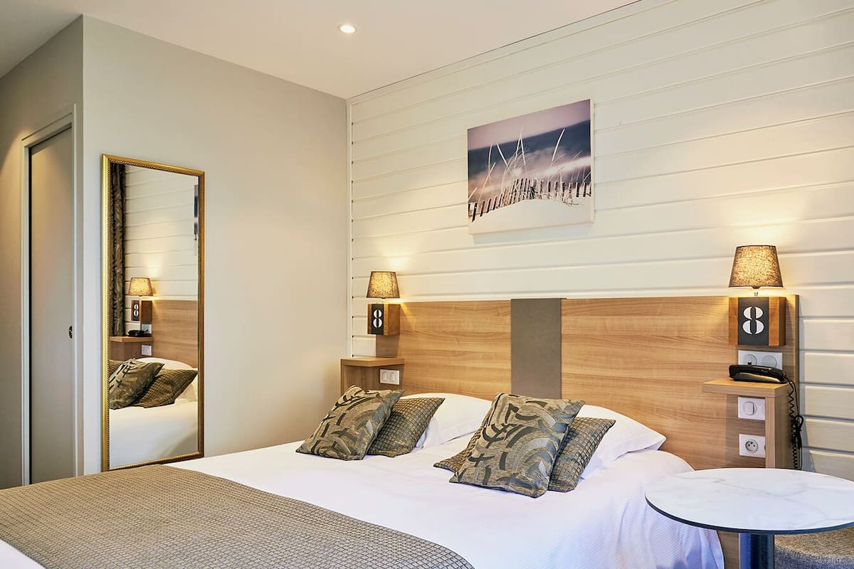 A hotel room with a bed, bedside lamps, a round table, a mirror, and a wall-mounted artwork.