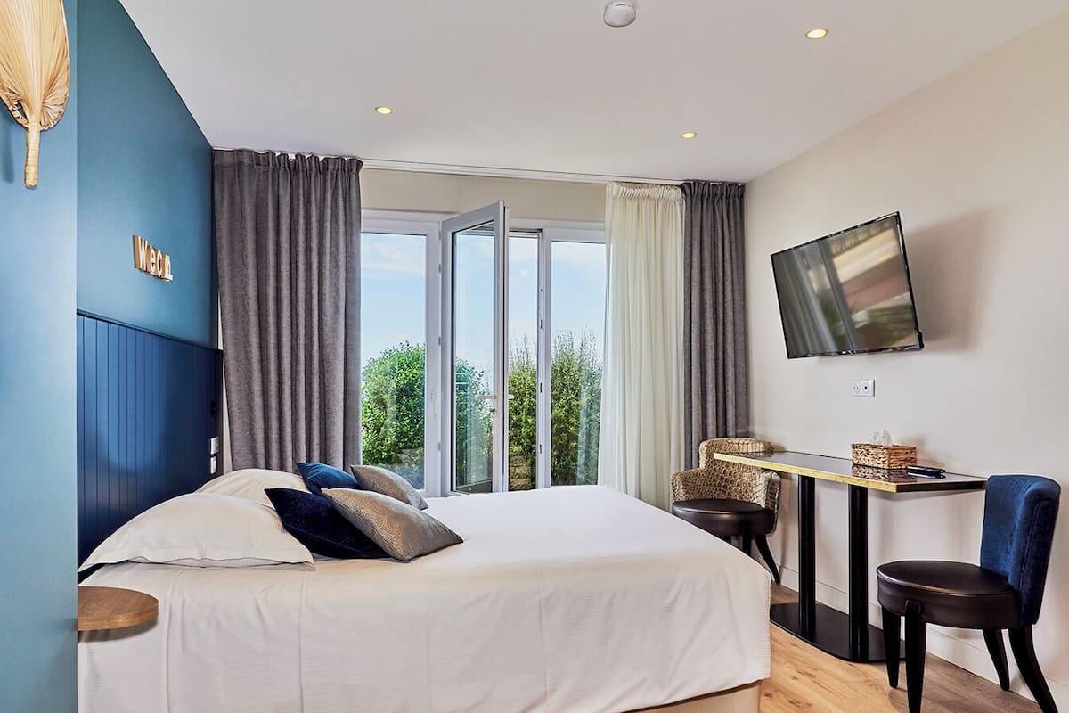 A bedroom with a bed, a desk, and a chair, a TV mounted on the wall, and a view of greenery outside.