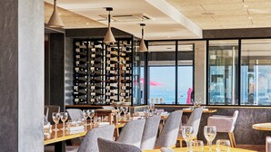 A restaurant with multiple tables and chairs, a wine rack, and a view of the sea through large windows.