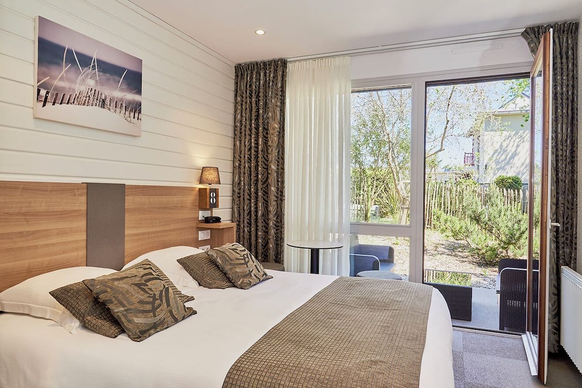 A hotel room with a large bed, a bedside table with a lamp, a view of the outdoors through a sliding glass door, and a painting on the wall.