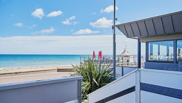 A beachside view with a clear sky, a sandy beach, and a building with large windows overlooking the ocean.