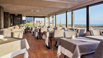 A restaurant with tables and chairs arranged for dining, large windows offering a view of the sea, and a modern interior design.