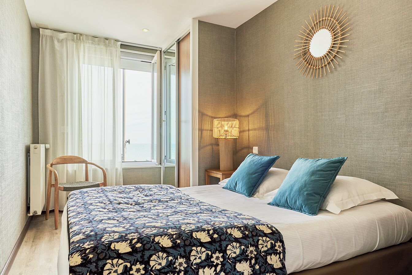 Quarto de hotel com uma cama grande, uma poltrona, um abajur e vista para o exterior.