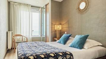 Quarto de hotel com uma cama grande, uma poltrona, um abajur e vista para o exterior.