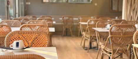 Interior de restaurante con sillas y mesas de mimbre, una máquina de café y obras de arte en las paredes.