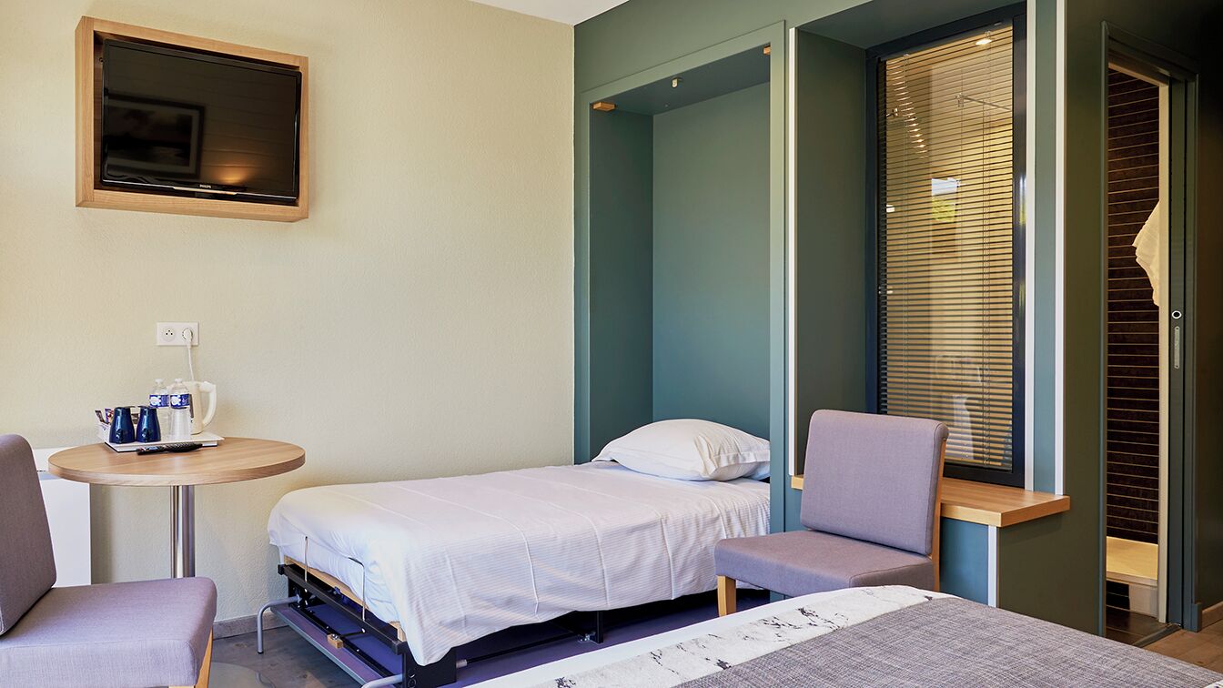 Habitación de hotel pequeña con una cama individual, una mesita con hervidor, una silla y una ventana con persianas.