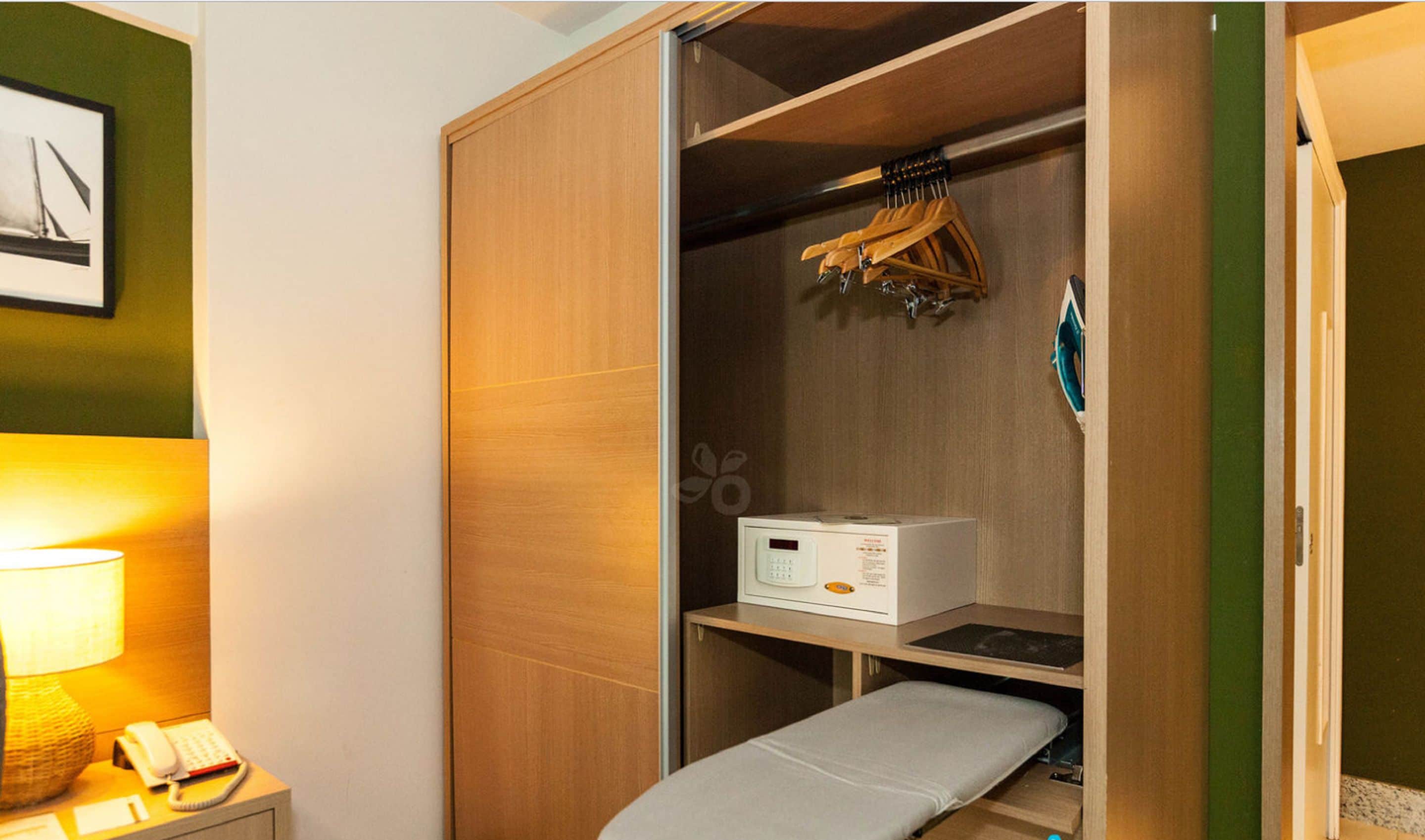 Una habitación compacta con un armario de madera, una mesita de noche pequeña con lámpara y una cama plegable.