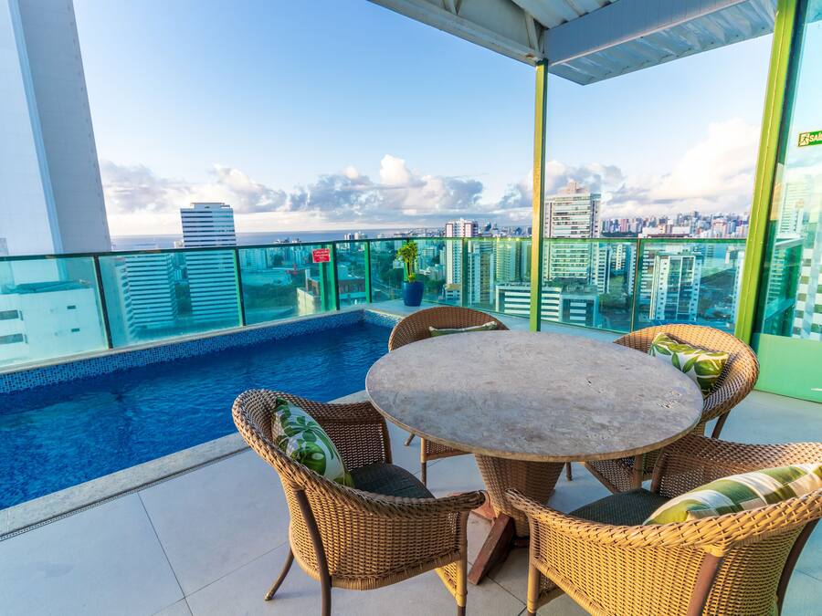 A rooftop pool area with wicker chairs and a table, overlooking a cityscape.