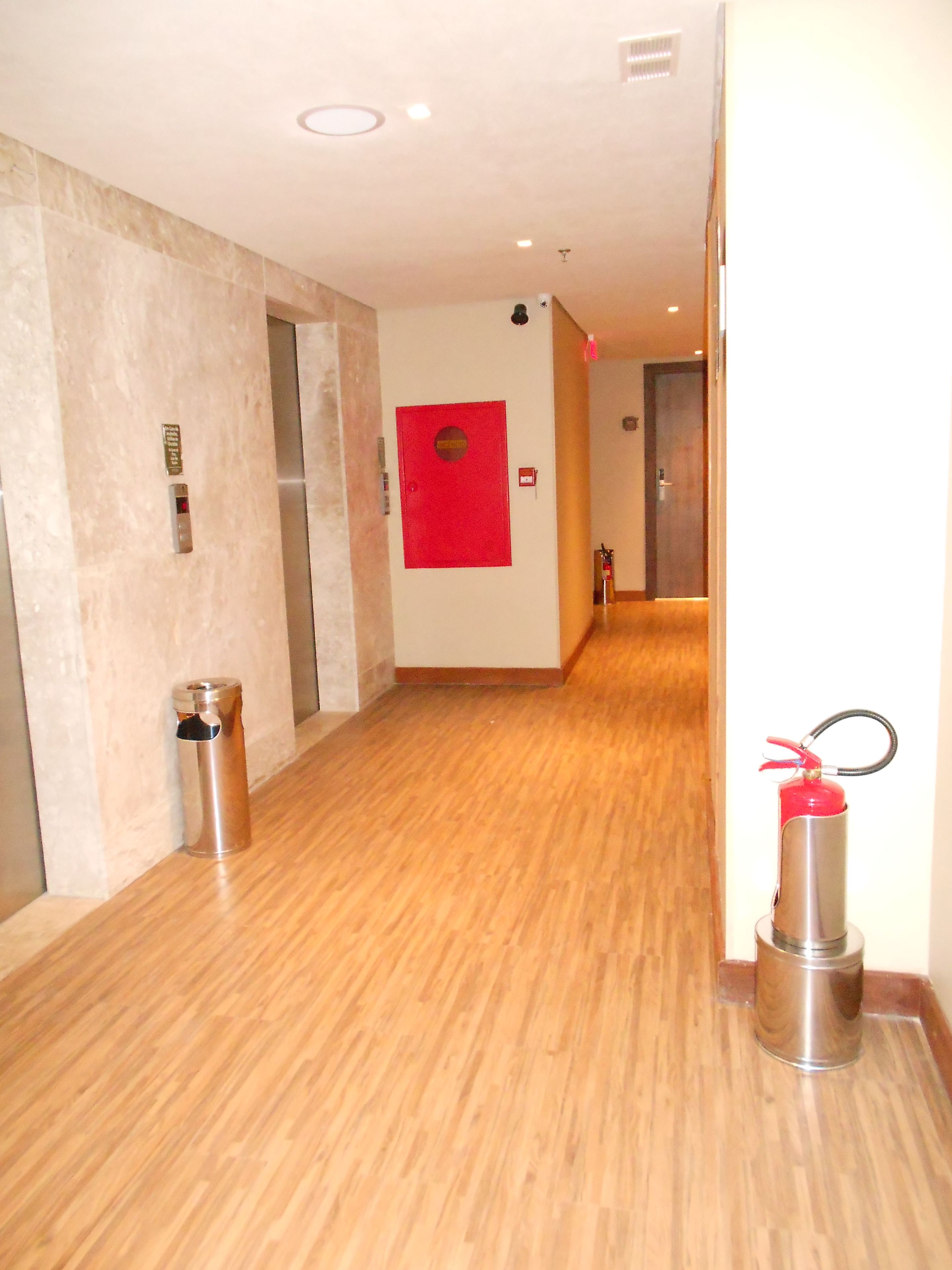A hallway with a fire extinguisher, a trash bin, and a red emergency exit sign.