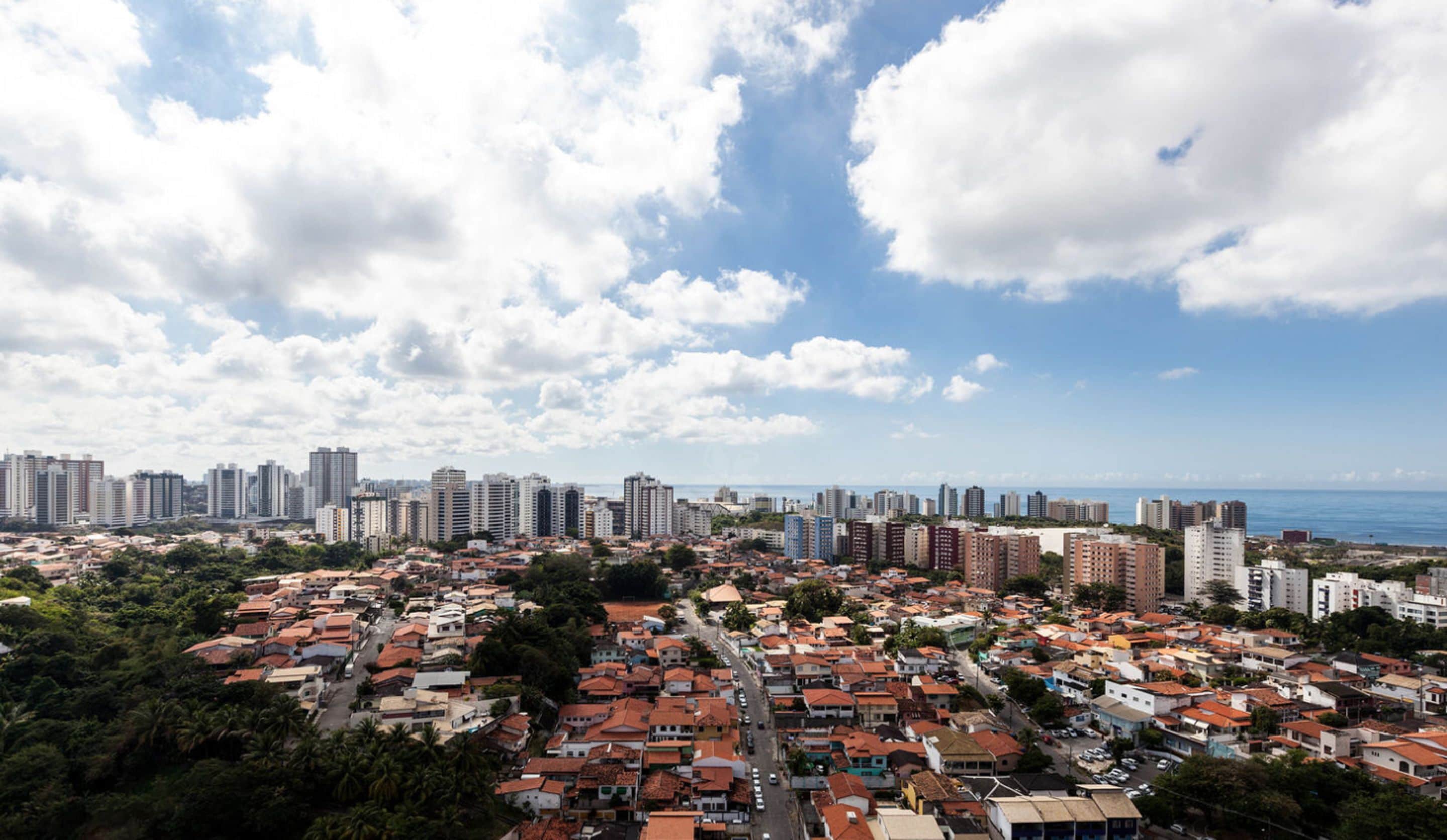A cityscape with a mix of high-rise buildings and low-rise residential areas, a clear sky, and a distant view of the sea.