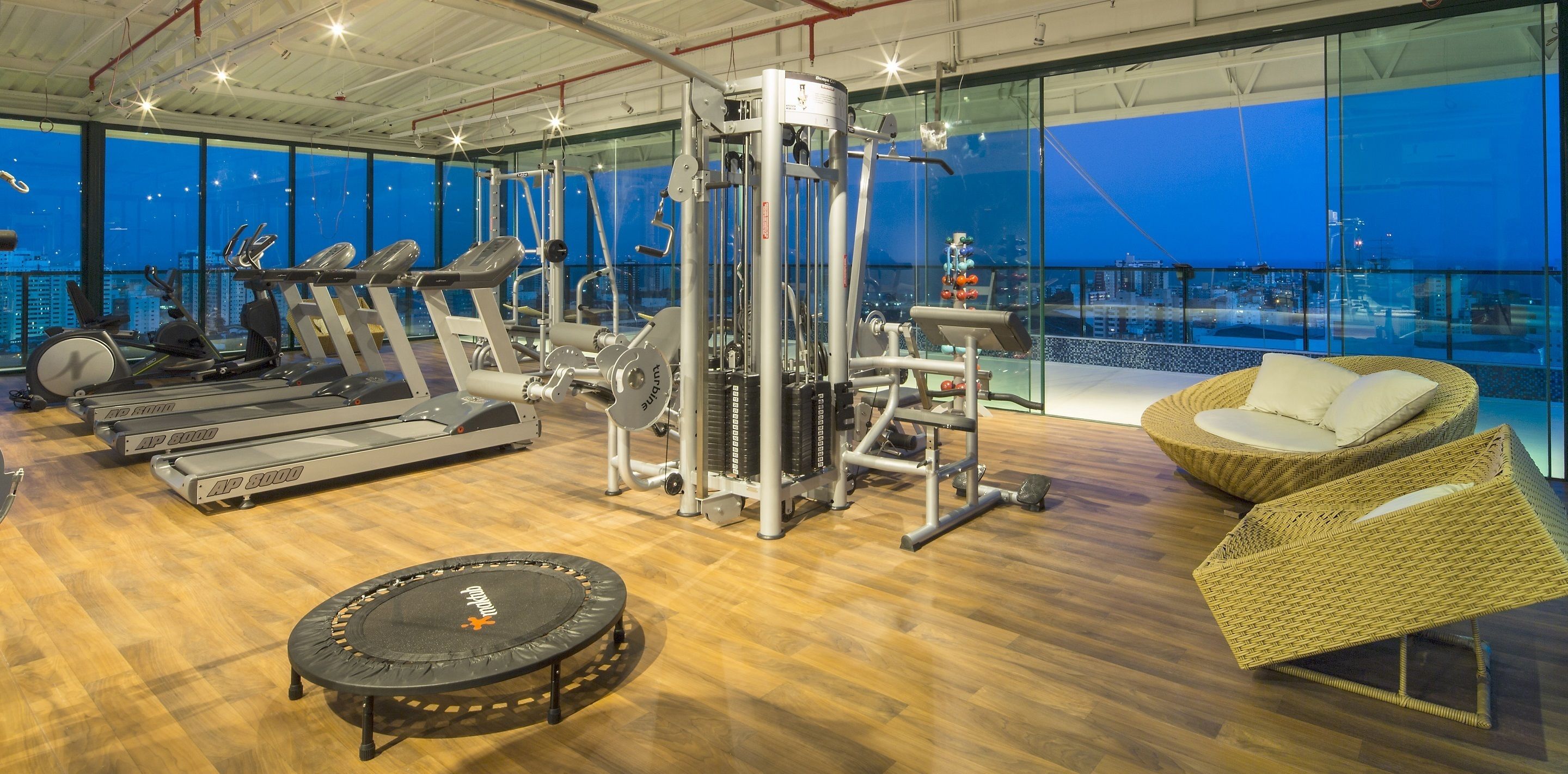 Une salle de sport équipée de matériel d’exercice, avec un trampoline et des meubles en osier, offrant une vue sur la ville au crépuscule.