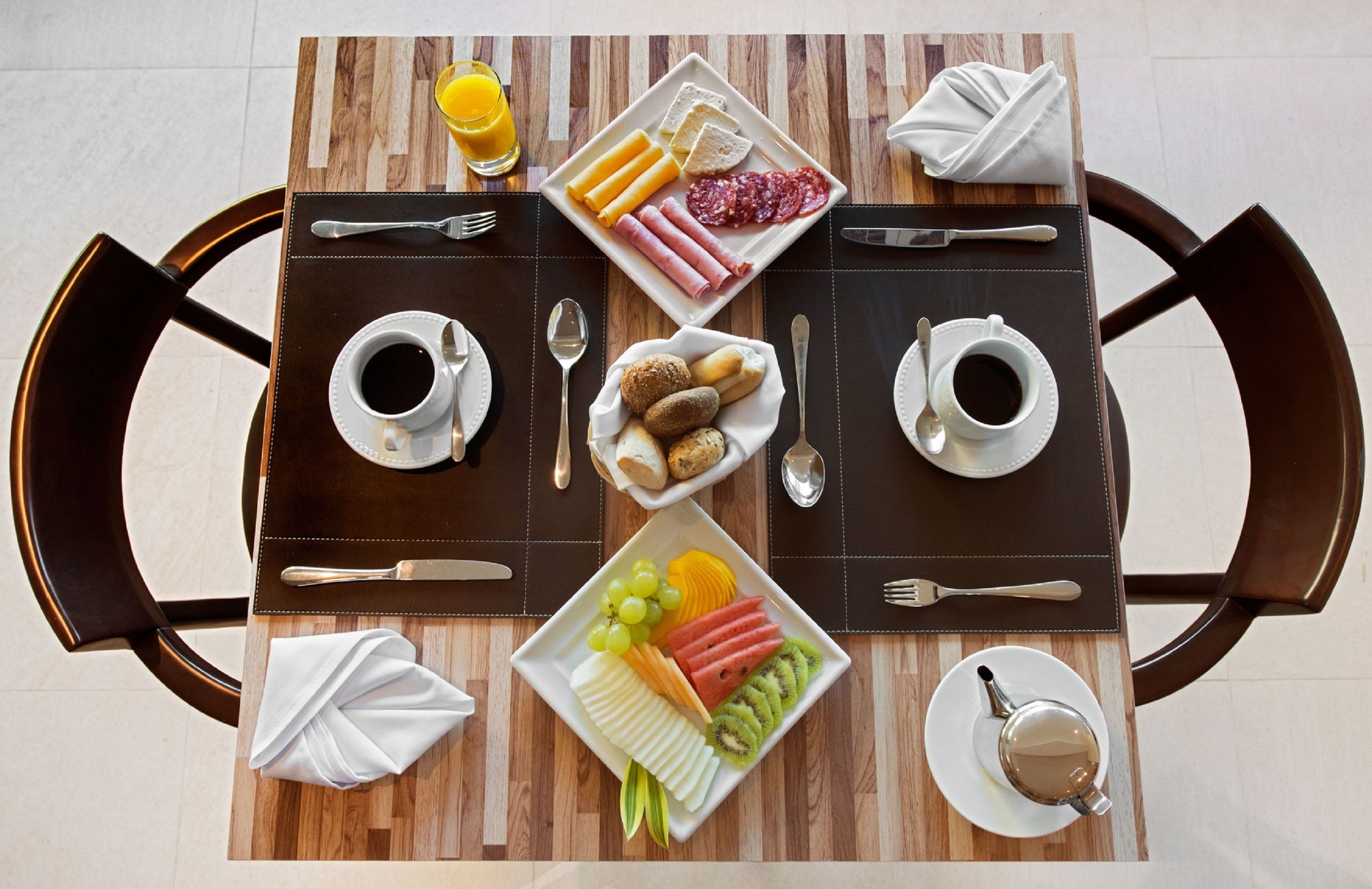 Ein Frühstückstisch mit Holzoberfläche, zwei Stühlen und einer Auswahl an Speisen wie Brot, Fleisch, Obst und Getränken.