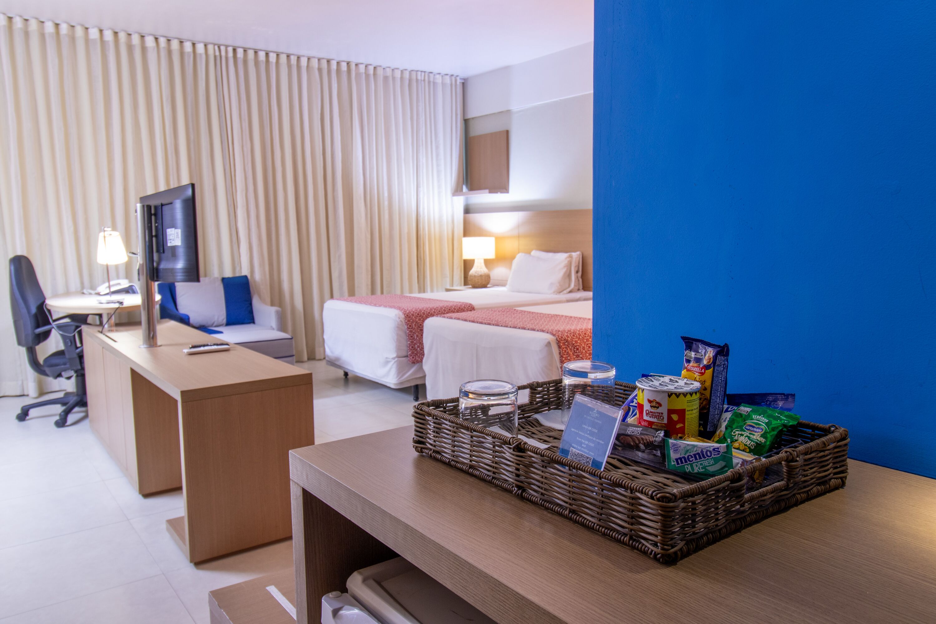 Habitación de hotel con escritorio, dos camas, televisor, lámpara y una cesta de mimbre con aperitivos.