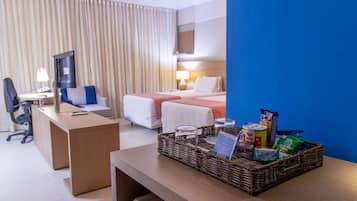 Habitación de hotel con escritorio, dos camas, televisor, lámpara y una cesta de mimbre con aperitivos.