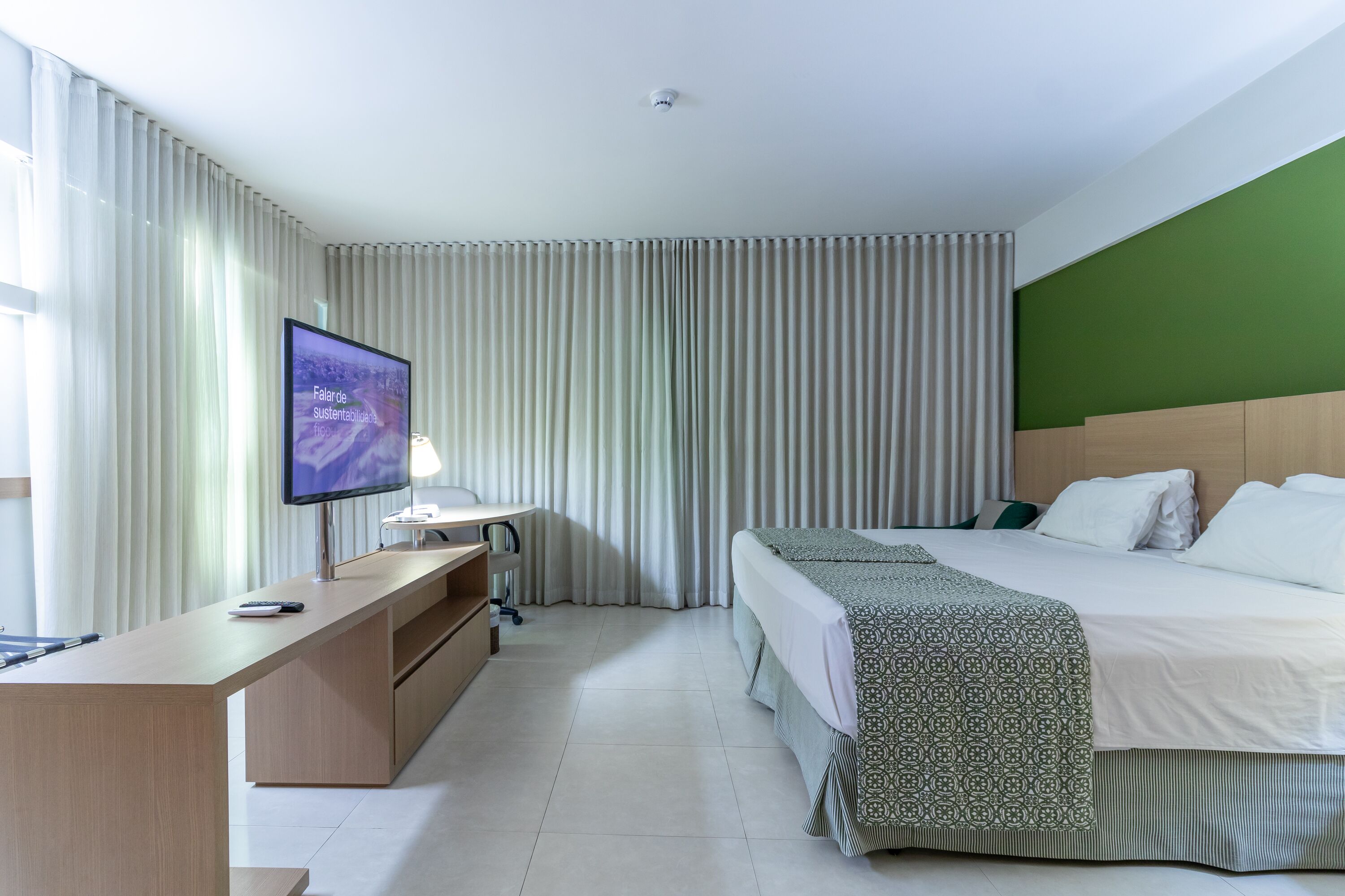 A hotel room with a bed, a desk, a television, and a green accent wall.