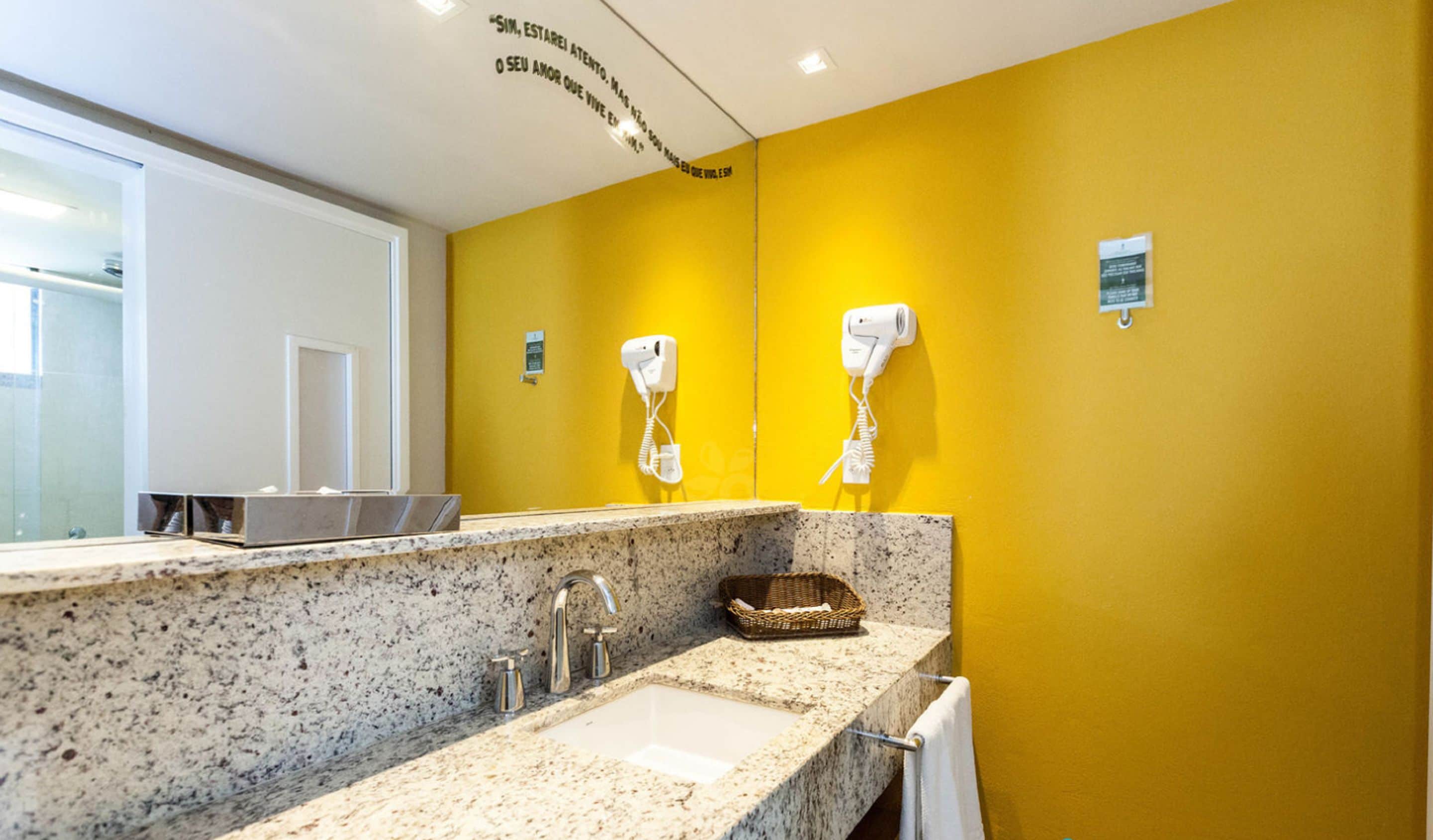 Casa de banho com bancada de granito, pia, espelho e dois secadores de cabelo fixados na parede.
