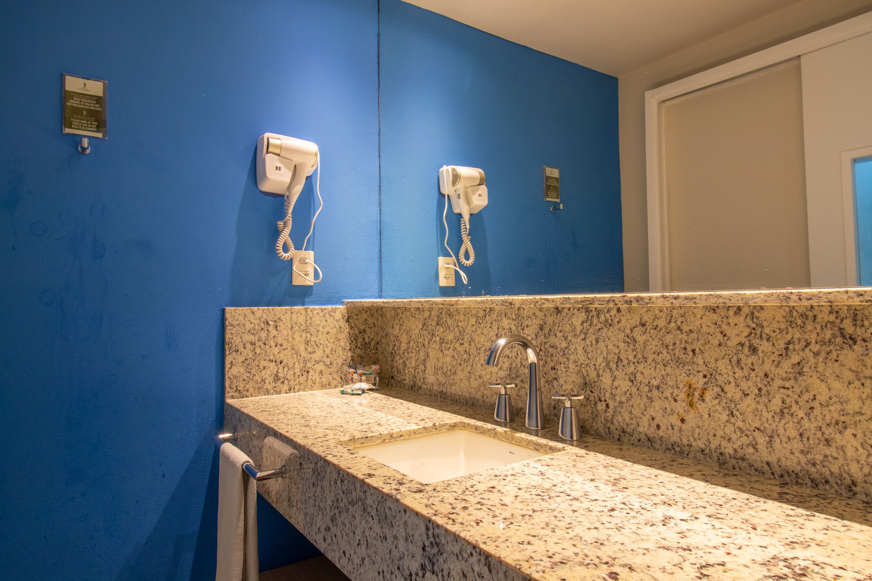 A bathroom with a granite countertop sink, two white hair dryers mounted on the wall, and a white door in the background.