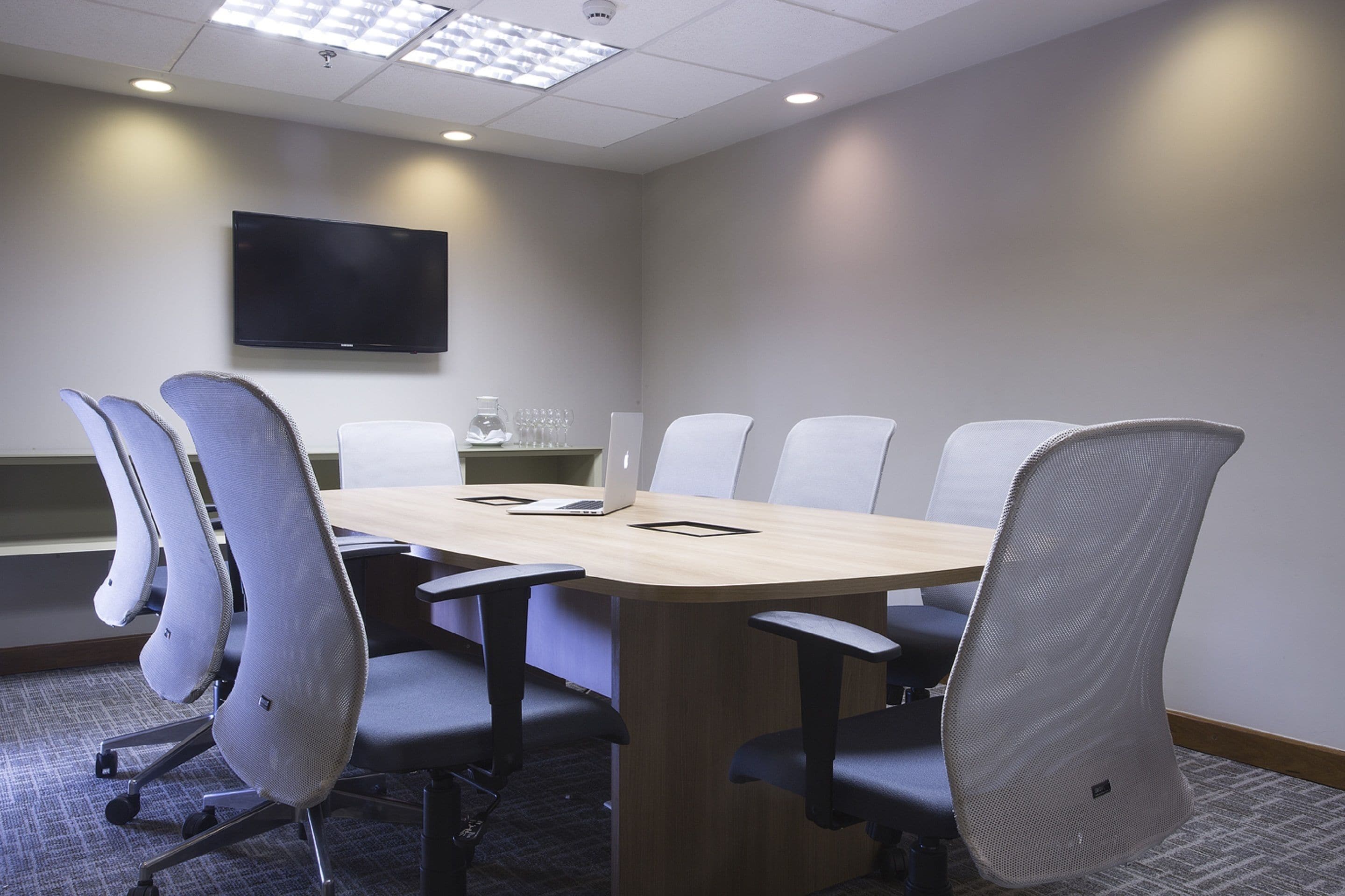 A conference room with a long table, six chairs, a flat-screen TV on the wall, and a coffee machine.
