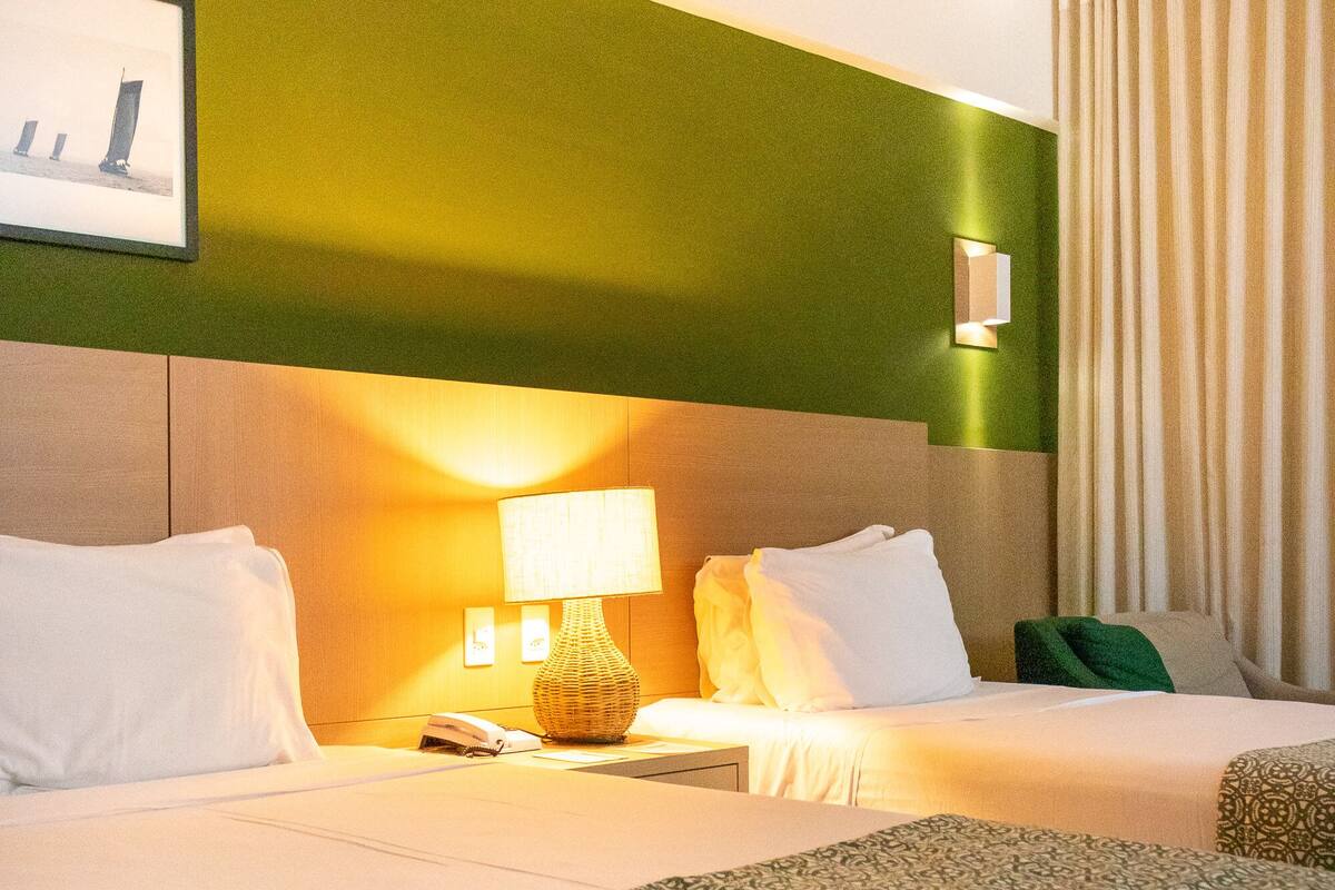 A hotel room with two beds, a green accent wall, a lamp, and a framed picture.