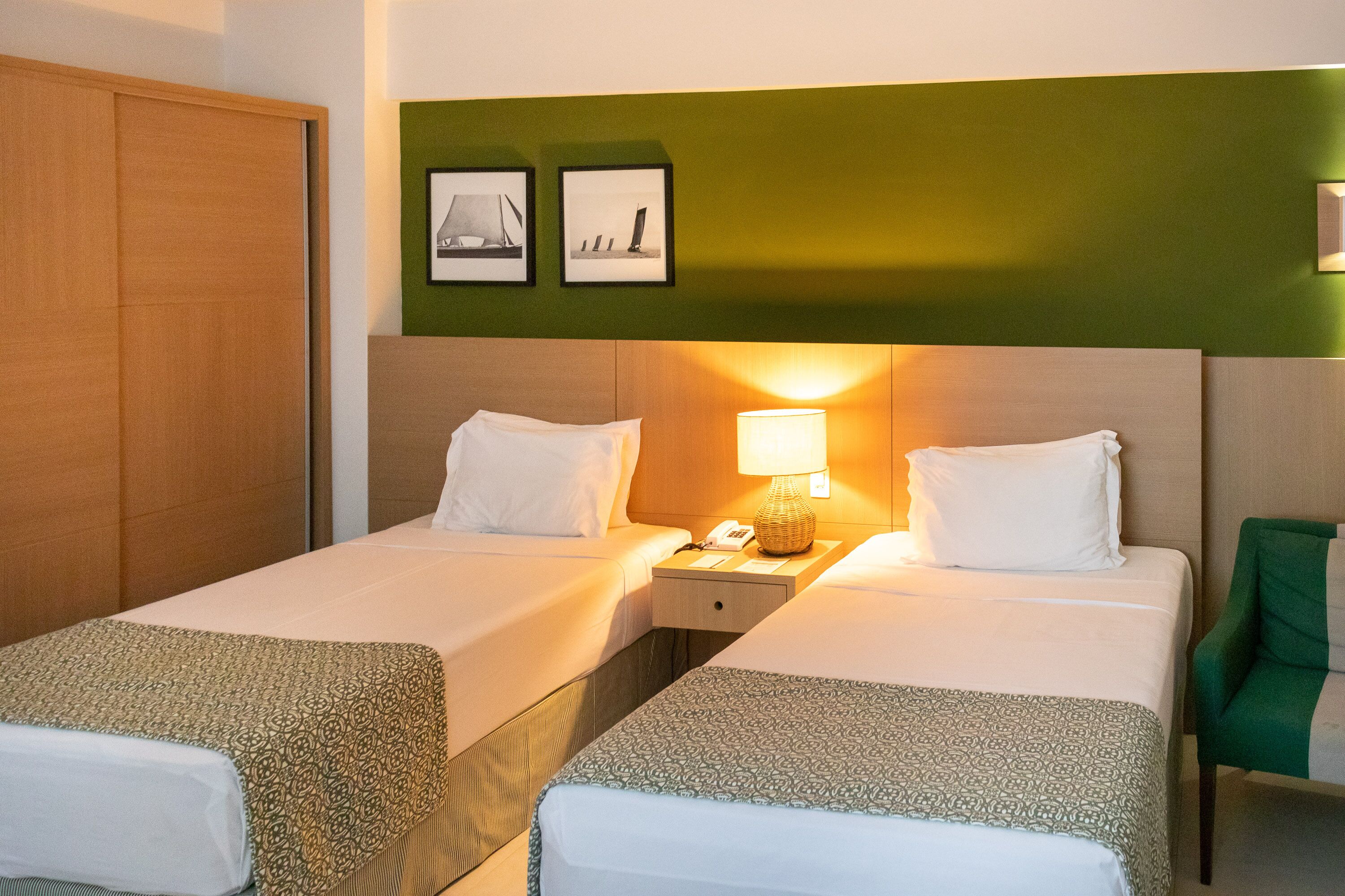 Une chambre d’hôtel avec deux lits, une table de chevet avec une lampe, un fauteuil vert et deux tableaux encadrés au mur.
