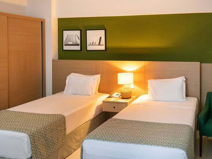 A hotel room with two beds, a nightstand with a lamp, a green chair, and two framed pictures on the wall.