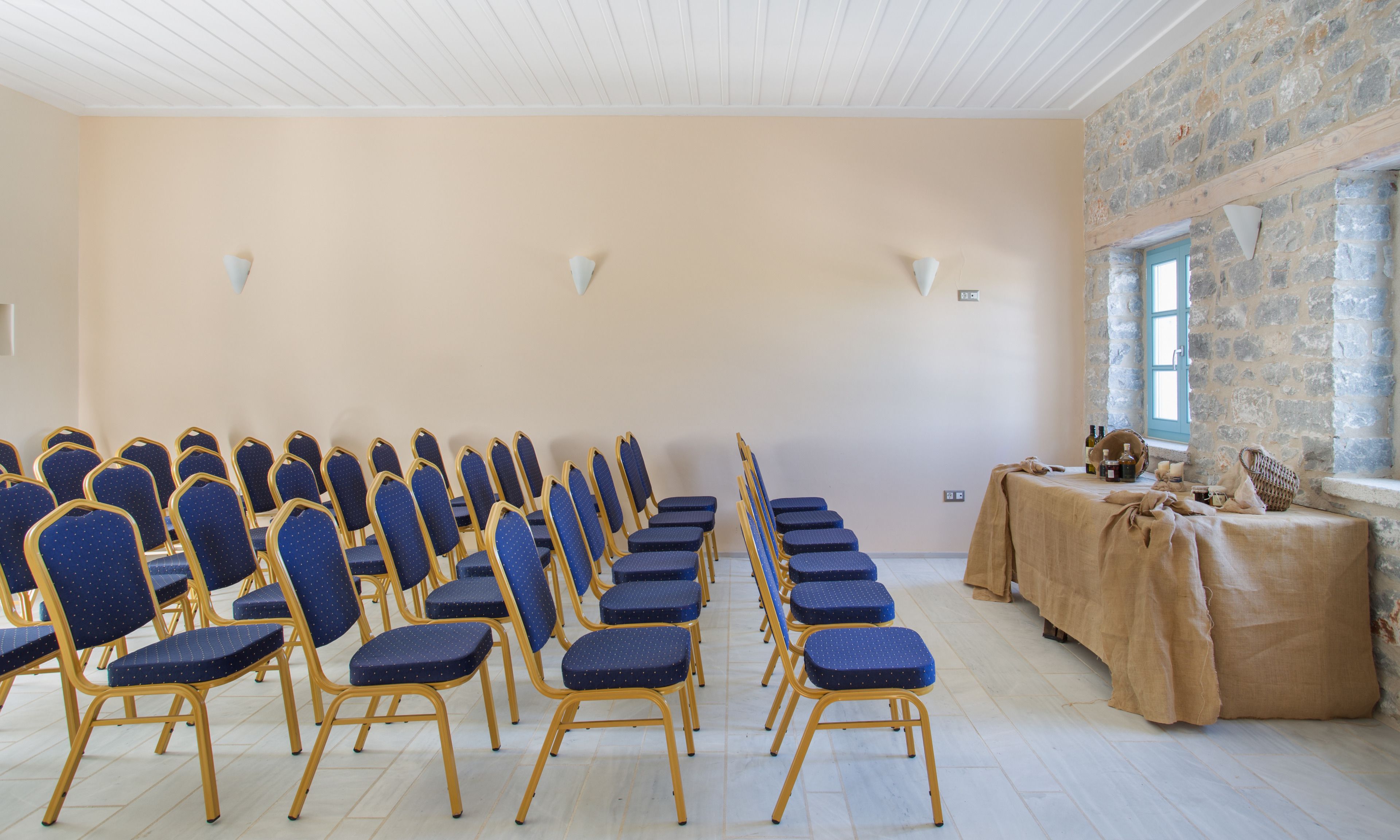 A conference room with rows of blue chairs, a table with a beige cloth, and a stone wall with a window.