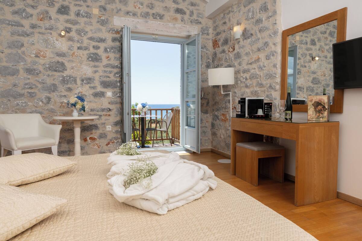 A room with a stone wall, a wooden desk, a white chair, a bed with white bedding, and a view of the sea through an open door.