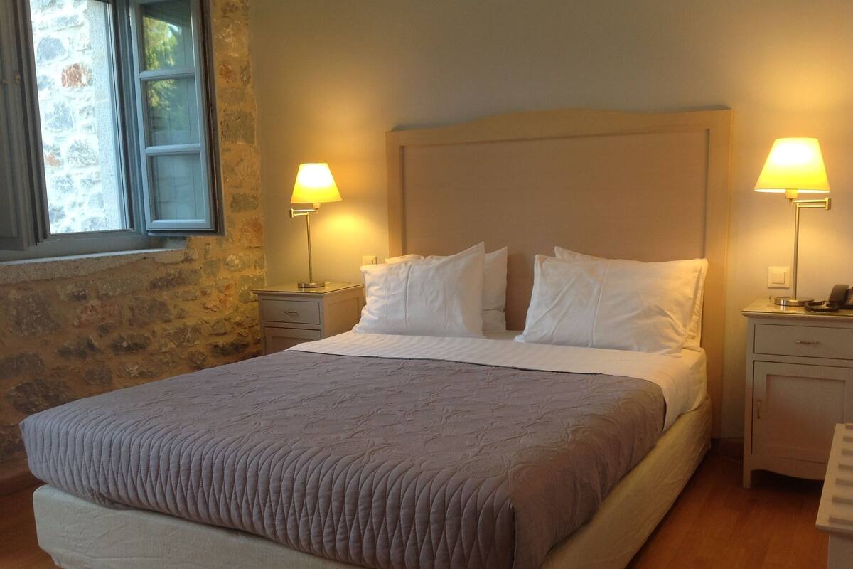A neatly made bed with a headboard, two bedside tables with lamps, and a stone wall to one side.