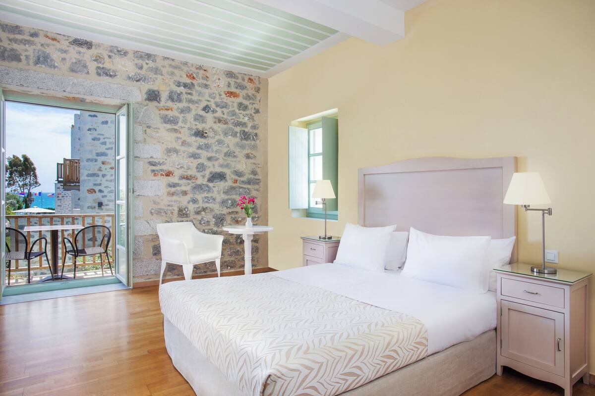 A bedroom with a large bed, a bedside table, a nightstand, a chair, a small table, and a stone wall.