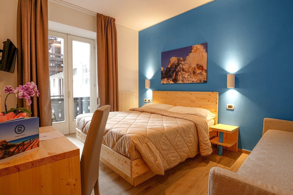 Een slaapkamer met een houten bed, een bureau met stoel, een raam met gordijnen, een schilderij aan de muur en uitzicht op buiten.