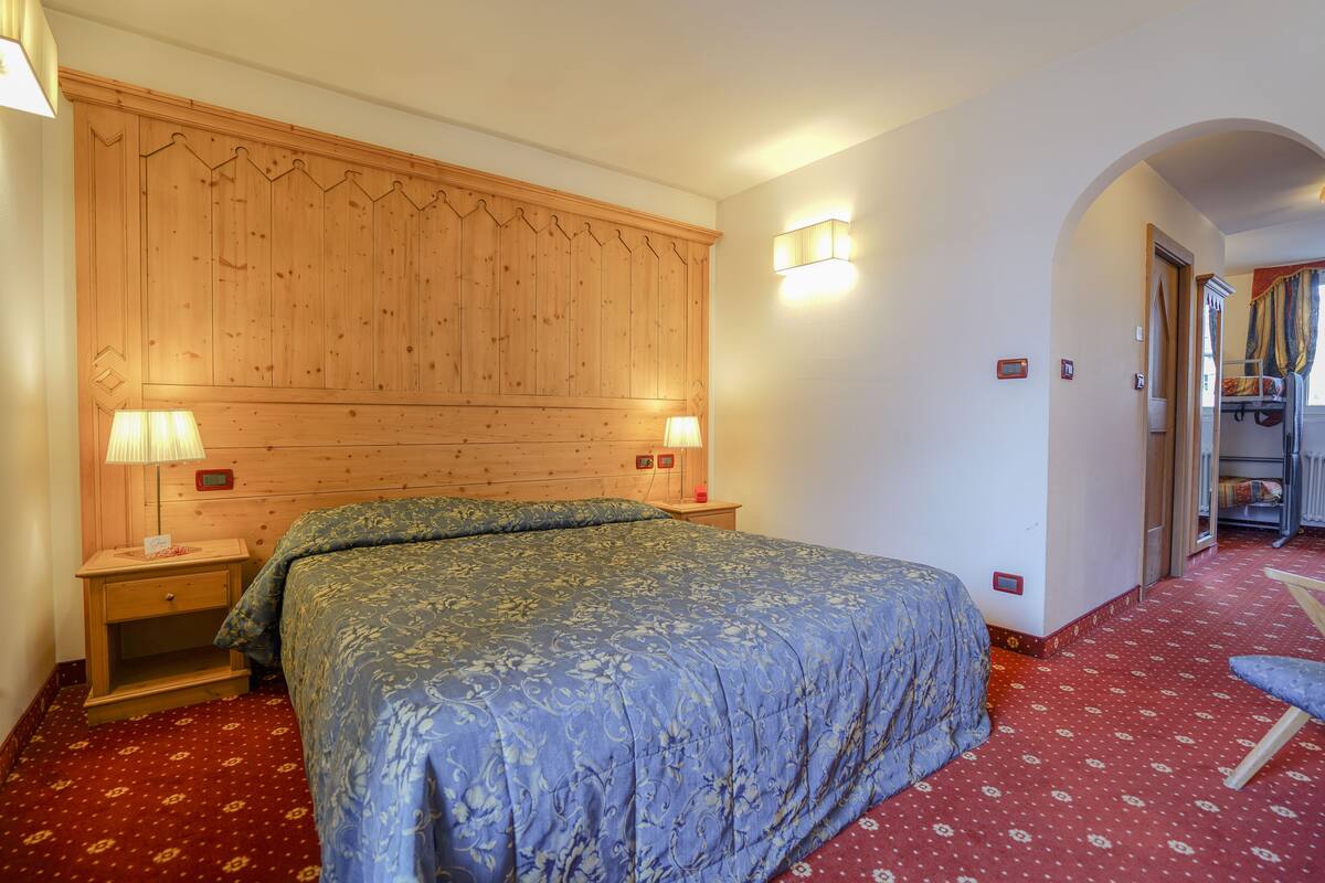 Een slaapkamer met een houten hoofdbord, twee bedlampen, een bed met een blauw bloemenbedover, een rood gestreept vloerkleed en een klein houten nachtkastje.