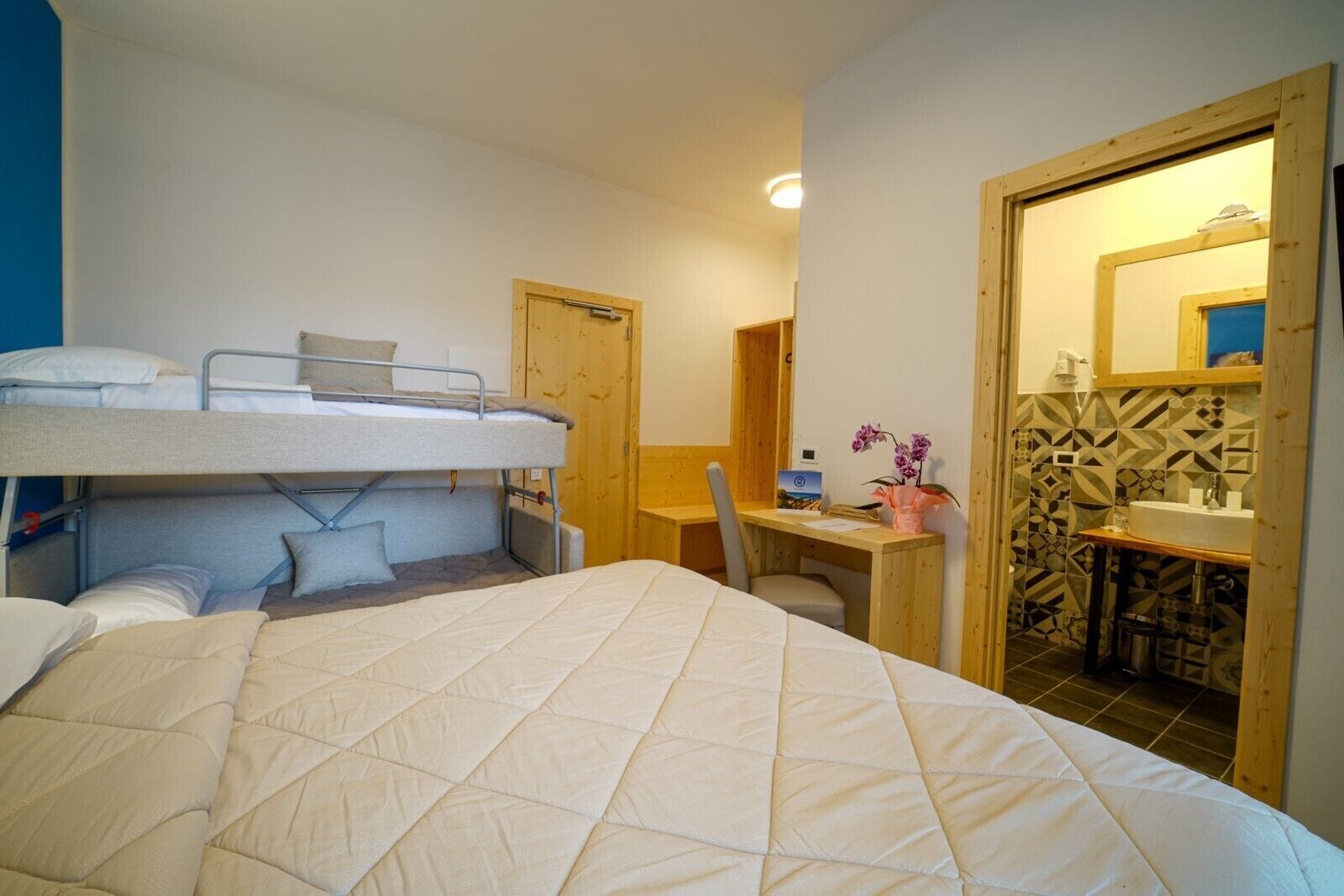 A dorm-style room with bunk beds, a desk, and a bathroom with a tiled backsplash.