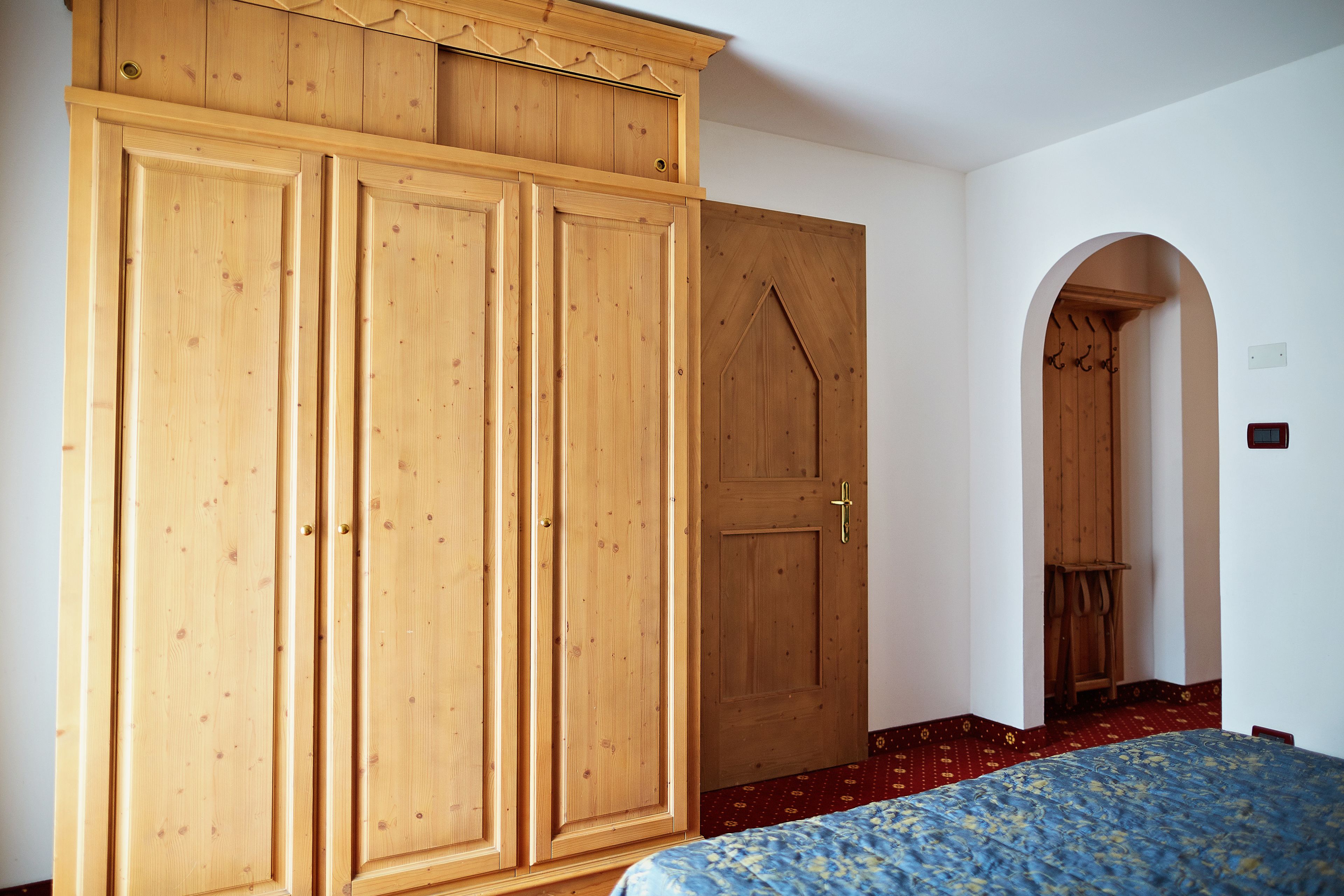 Une chambre à coucher avec une grande armoire en bois, un lit recouvert d’une couvre-lit à motifs et une porte en bois dotée d’une poignée.
