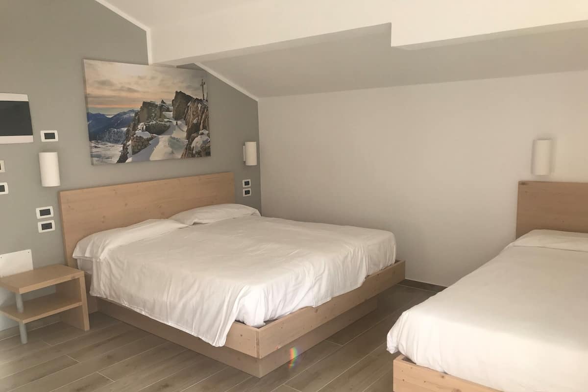 Een moderne slaapkamer met een groot bed, een kleiner bed, een houten nachtkastje, een bijzettafeltje en een schilderij van een bergachtig landschap.