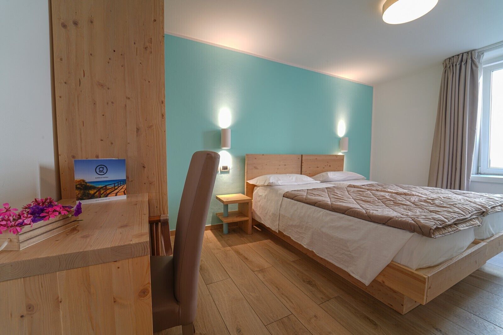 Une chambre d’hôtel avec un lit en bois, un bureau avec une chaise, une table de chevet et une lampe fixée au mur.