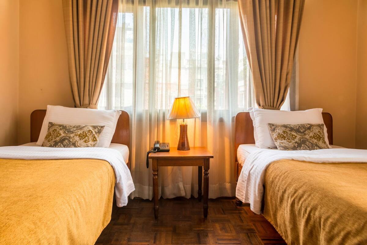 A hotel room with two beds, a nightstand with a lamp, and sheer curtains.