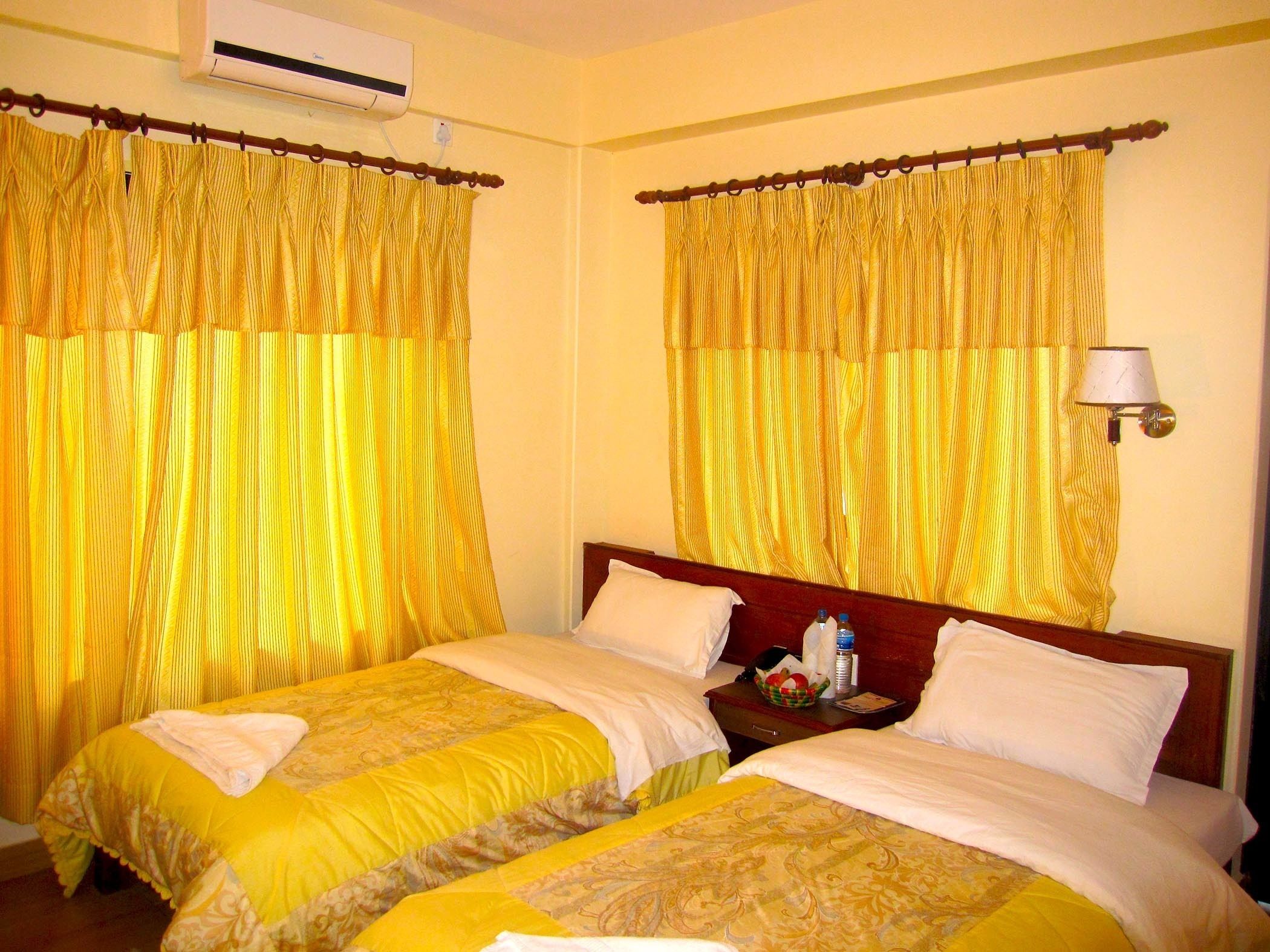 A hotel room with two beds, yellow curtains, a bedside lamp, and a small table with a basket and bottles.