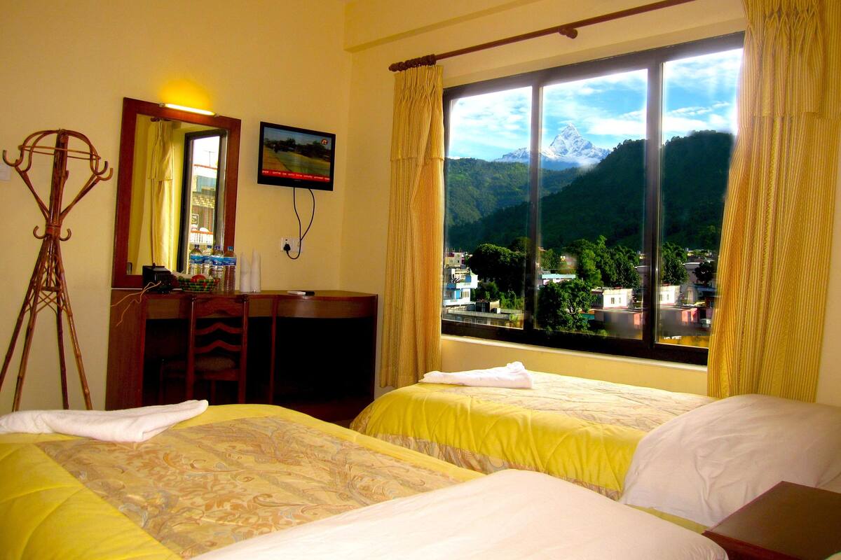 A hotel room with two beds, a desk, a chair, a mirror, and a window with a mountain view.