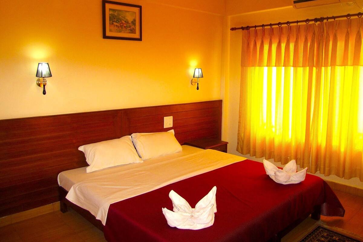 A hotel room with a wooden headboard, two bedside lamps, a painting on the wall, and a bed with a red bedspread and white pillows.