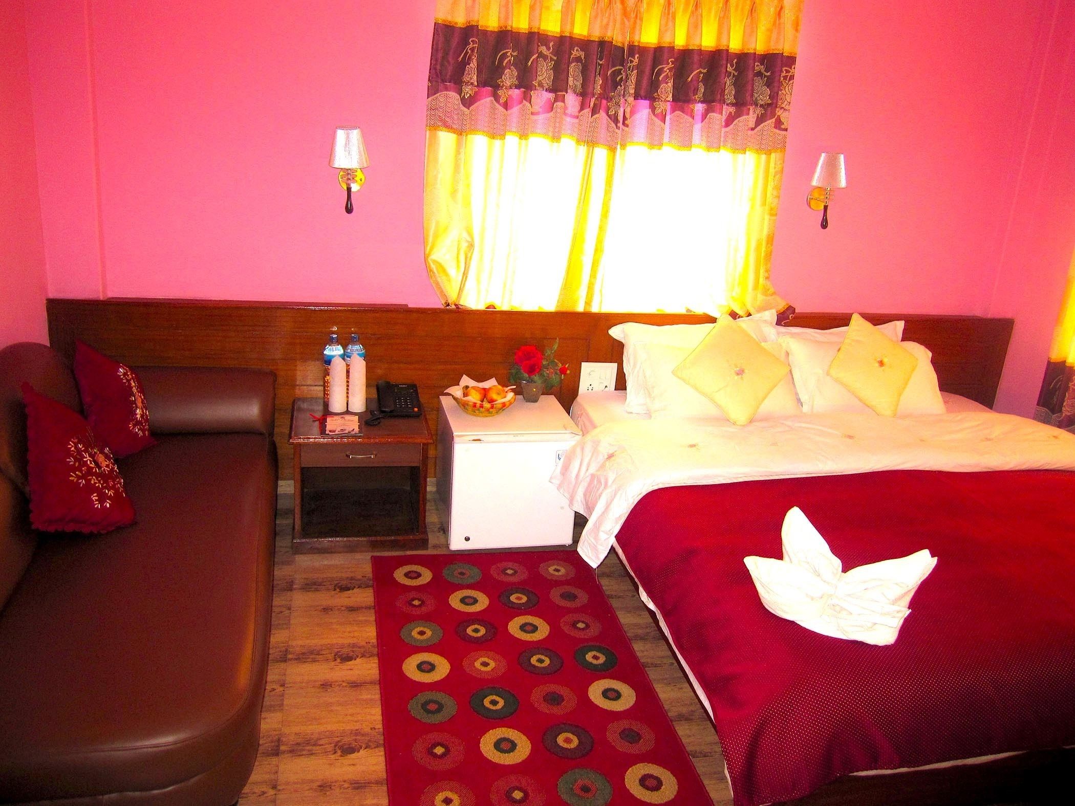 A hotel room with a bed, a sofa, a nightstand with a lamp, and a small table with a vase and a basket of fruit.